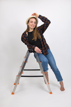 Beautiful barefoot blue-eyed smiling girl with long blond hair wearing a hat expresses emotions of satisfaction while posing while sitting on a folding aluminum ladder. Close-up on a white background.の写真素材