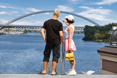 A family, a mother with an adult son, walks along the embankment against the backdrop of a bridge over the river on a sunny summer day.の写真素材