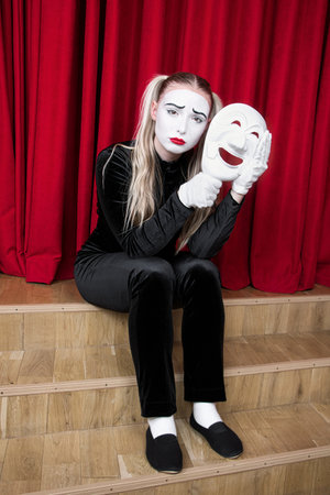 Portrait of a sad mime girl with a mask in her hands sitting on a theater stage.の写真素材