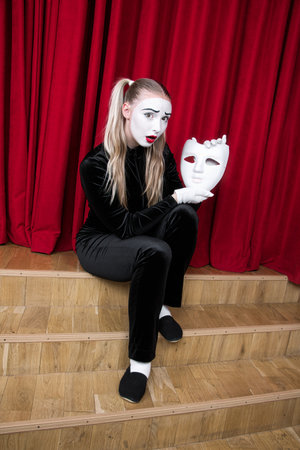 Portrait of a mime girl with a mask in her hands, sitting on a theater stage.