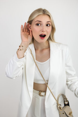 Blonde teenager in white suit puts hand to ear to express emotions of eavesdropping. Close-up on light background.の写真素材