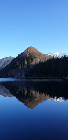 Beautiful Siberian winter landscape with mountain reflection on calm water of the Snezhnaya River. Clear blue sky and forested slopes create a peaceful natural scene.の写真素材