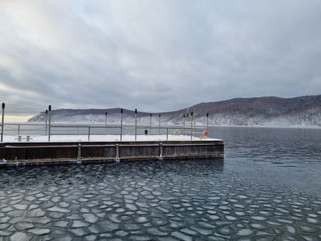 Beautiful winter view of a frozen pier surrounded by ice formations on Lake Baikal, Listvyanka, Russia. Snowy hills, cold mist, and calm water create a peaceful Siberian landscape.の写真素材