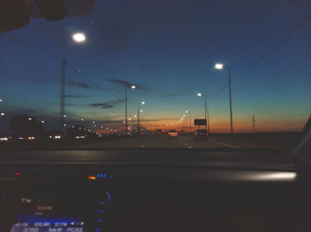 View through the windshield of a car driving along a highway during sunset. The streetlights and vehicles ahead create a calm yet dynamic travel atmosphereの写真素材