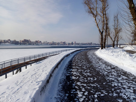Snowy embankment and frozen river in Irkutsk, Russia, on a bright sunny winter day. Peaceful Siberian landscape with bare trees, clear blue sky, and quiet city viewの写真素材