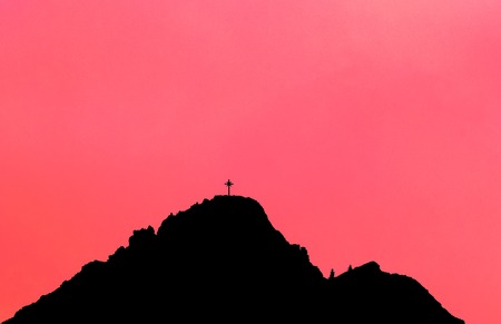 Black silhouette of mountain peak with summit cross. Pink background.の写真素材