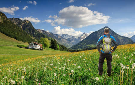 Hiker with Backpack standing on Flower meadow. Snow covered mountains and traditional house. Bavaria, Alps, Oberstdorf, Allgau, Germany.の写真素材