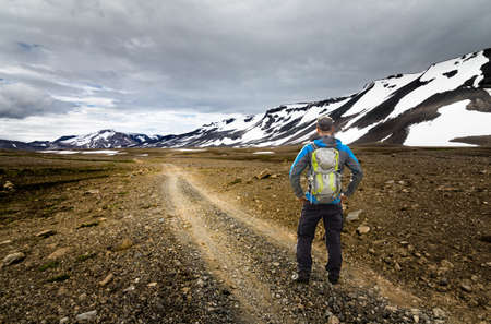 Young Man with Backpack hiking on dirt road trail in arctic desert and snow mountains of Iceland. Kaldidalur, Thorisjokull, Iceland. View to Thorisdalur.の写真素材