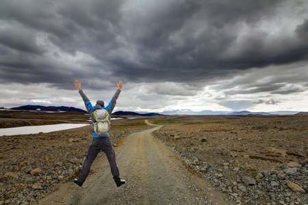 Happy Sucessful Winning Hiking young Man Jump with Backpack and raised arms on Trail road. Hiking in arctic desert of Iceland on Road F550. Kaldidalur, Iceland.の写真素材