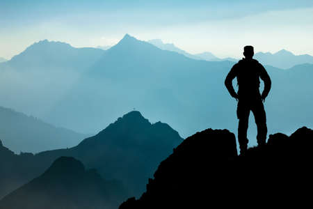 One Man reaching summit after climbing and hiking enjoying freedom and looking towards mountains silhouettes panorama during sunrise.の写真素材