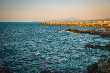 Experience the enchantment of twilight on a rocky beach in Rethymno. Against the backdrop of majestic mountains and with a ship gliding across the sea, the scene is bathed in a warm golden hue from the setting sunの写真素材