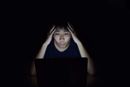 Asian man using laptop in the black dark room and show surprise something on his face