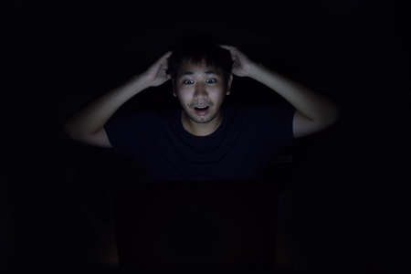 Asian man using laptop in the black dark room and show surprise something on his faceの写真素材