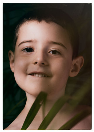 The boy looks at the camera, with palm leaves in the foreground. Childrens portrait through the...