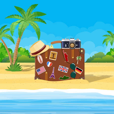 Vintage old travel suitcase on beach. Leather retro bag with stickers. Hat, photo camera,...