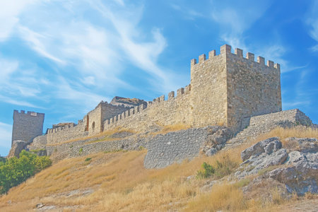 A historic fortress stands atop a hillside, featuring stone walls and towers against a bright sky.の素材