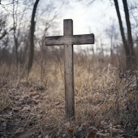 A solitary wooden cross rises from the ground in a grassy area, surrounded by dried leaves and sparse vegetation, as the light dims just before evening.の素材