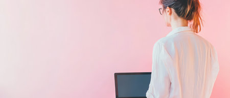 A woman stands in a light colored space focused on her laptop while working creatively. The soft pink background adds to the artistic atmosphere.の素材