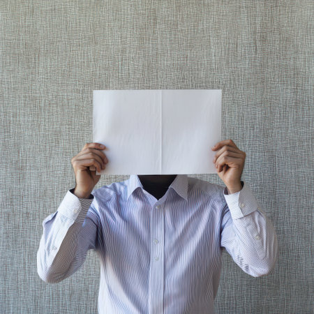 A person stands against a textured background holding a blank sheet of paper in front of their face creating a sense of anonymity and curiosity.の素材