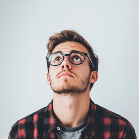 A young man wearing glasses and a plaid shirt looks thoughtfully upward with a curious expression in a simple indoor setting.の素材