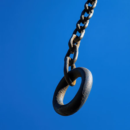 A metal chain link hangs from above illuminated by sunlight against a bright blue sky with no clouds creating a vivid contrast.の素材