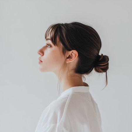 A young woman stands in profile against a plain background showcasing her elegant bun hairstyle and thoughtful expression in soft lighting.の素材
