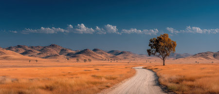 A winding dirt road cuts through golden grasslands leading to a solitary tree against a backdrop of rolling hills and blue sky.の素材