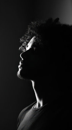 A young man gazes upwards with a thoughtful expression highlighted by contrasting shadows on his face and hair in a minimalist setting.の素材