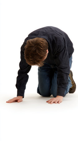 A man dressed in a black striped shirt and jeans kneels on the floor appearing deep in thought or reflection in a minimalist setting.の素材