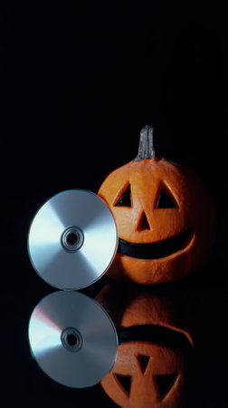 A carved pumpkin with a smiling face sits beside a shiny CD. The scene is dimly lit evoking a festive Halloween atmosphere.の素材