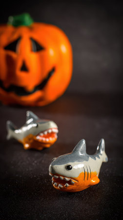 Two small shark toys sit on a dark surface with a carved Halloween pumpkin in the background creating a festive and playful atmosphere.の素材