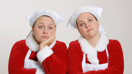 Two women dressed in red holiday costumes sit side by side showing expressions of boredom and disappointment while celebrating the festive season.の素材