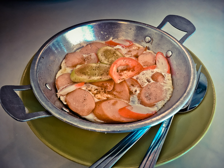 Fried eggs in pan on dish with hot-dog, tomato, vegetable and sauce on tableの写真素材