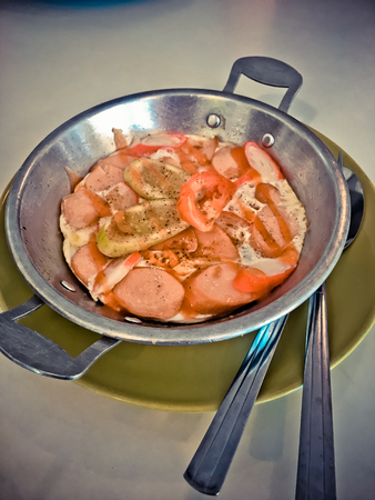 Fried eggs in pan on dish with hot-dog, tomato, vegetable and sauce on tableの写真素材