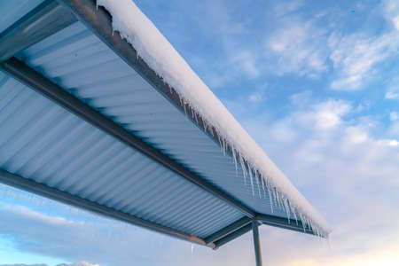 Snowy roof with icicles against cloudy blue skyの写真素材