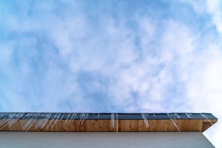 Roof with sharp icicles against cloudy blue sky. Looking up at the eaves of a roof with sharp...
