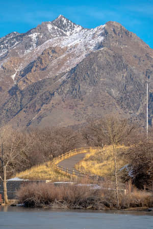 Lake road and mountain at Highland Glen Park Utah. Sunny day at Highland Glen Park with view of a fenced road beyond the calm lake. A rugged mountain against blue sky can be seen in the distance.の写真素材