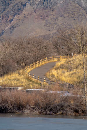 Narrow curving road with lake and mountain view. A narrow curving road lined with wooden at Highland Glen Park, Utah. The road has a view of a calm lake and immense mountain on a sunny day.の写真素材