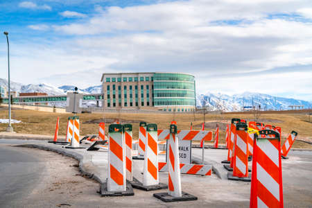 Sidewalk Closed sign and road safety poles on a paved road under repair. In the background is a modern building with skybridge and snowy mountain under a cloudy blue sky.の写真素材