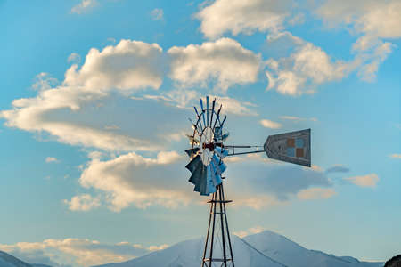 Windpump with a snow covered mountain and cloudy blue sky in the background. This multi-bladed steel...