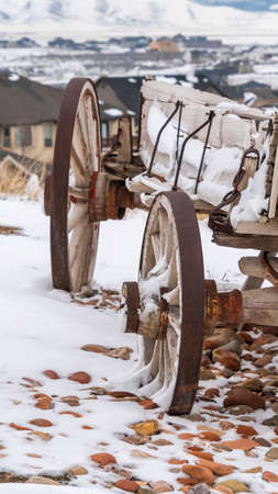Clear Vertical Weathered wooden wagon with chains and rusty wheel viewed in winter. The wagon is on a snowy hill overlooking homes and snow covered mountain.の写真素材