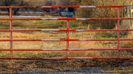 Clear Panorama No Trespassing sign on the gate of a private property viewed on a sunny day. A grassy terrain with trees against cloudy sky can be seen behind the weathered red gate.の写真素材