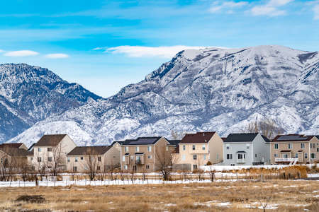 Row of beautiful homes against a rugged mountain dusted with snow in winter. A tranquil blue sky with white puffy clouds can be seen in the background.の写真素材