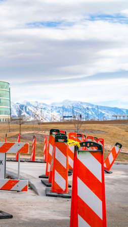 Vertical Sidewalk Closed sign and road safety poles on a paved road under repair. In the background is a modern building with skybridge and snowy mountain under a cloudy blue sky.の写真素材