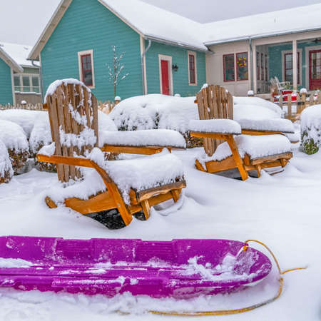 Square Purple sled and wooden chairs surrounded by snow during winter in Daybreak. Snow topped shrubs, fence, and home can be seen in the background.の写真素材