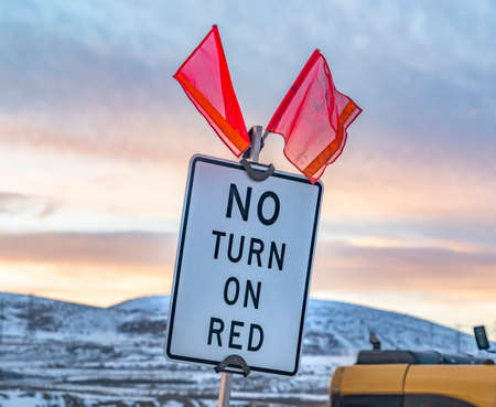 Close up of a No Turn On Red sign with red flags on top. In the background is a yellow vehicle and snow covered mountain under a cloudy sky in winter.の写真素材