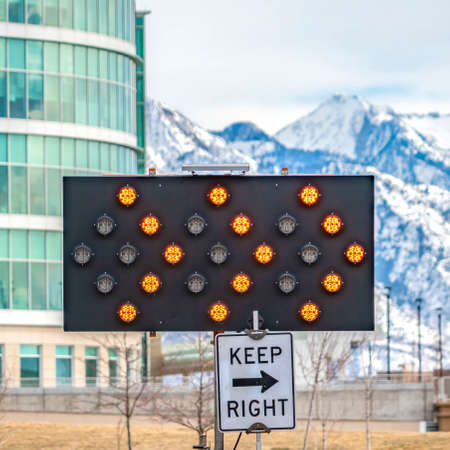 Clear Square Keep Right sign underneath an arrow board surrounded by road safety poles. A modern glass building and snow capped mountain against cloudy sky can be seen in the background.の写真素材