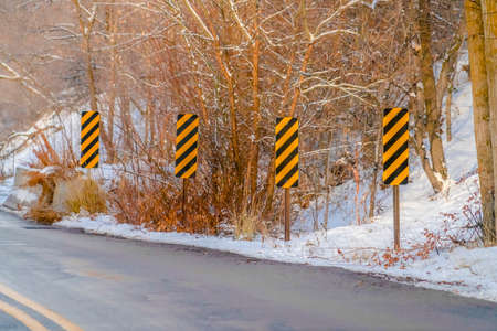 Mountain road and road signs viewed in winter. Yellow and black diagonal stripes road sign can be...