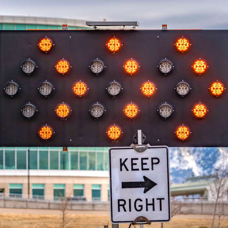 Square Close up of an arrow board and Keep Right sign against a modern building. A rugged snow...