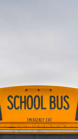Vertical Rear of a yellow school bus with signal lights and emergency exit window. A sky filled with clouds and snowy landscape in winter can be seen in the background.の写真素材